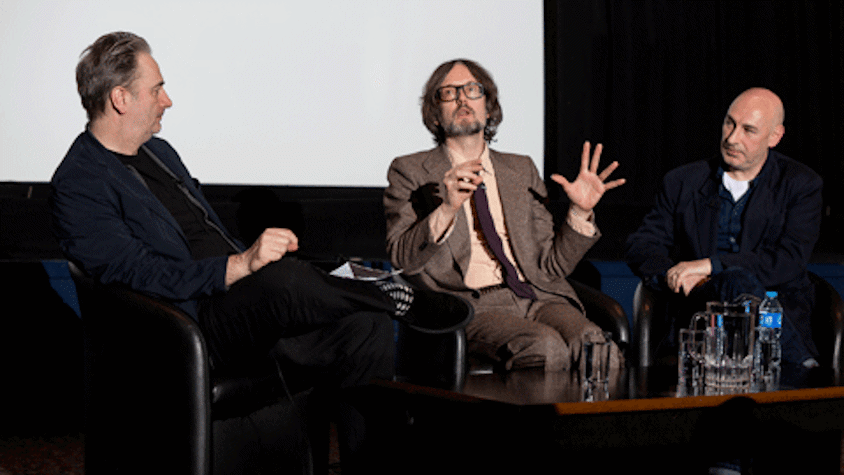 Three men are seated on chairs, there is a large projector screen behind them and a table with water glasses and a pitcher placed in front of them. The three men are seated in a way that they are all facing the front. The man in the middle is wearing a suit and is speaking with his hands open in expression. The man on either side of the speaker have their bodies and faces turned in his direction.