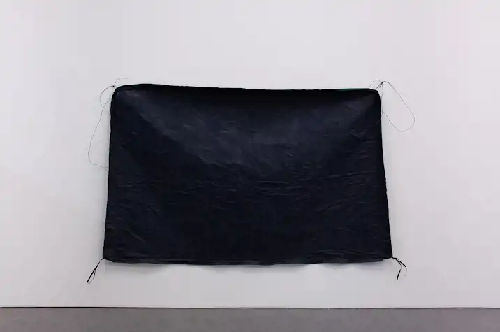 A piece of black waterproof-looking fabric, in a rectangle with rounded corners and fabric and cord tabs at each corner hangs on a white wall.