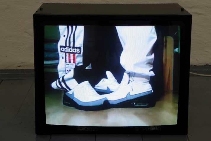 TV monitor on the floor showing the feet and lower legs of two people standing facing each other. they both wear nike trainers and tracksuit bottoms