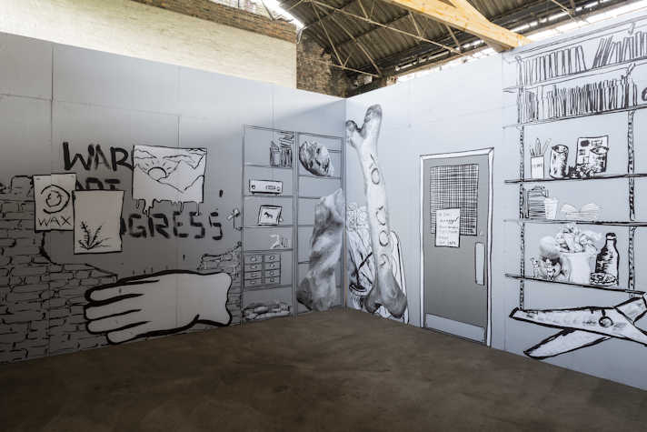 We can see two walls intersecting in a corner, both with drawn black and white murals. To the left we can see a message of "war not progress" with several posters in front of it. Underneath it, there is a giant hand and to the right some shelves with several objects. We can see on the wall on the right the giant bone that reads "to dog" and a door. To the right we can see more shelves with books and other objects. Below the shelves there are giant scissors.