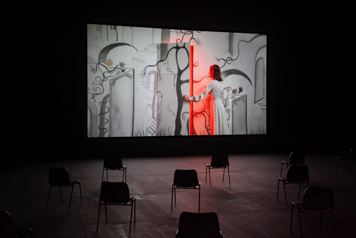 Image of a film being screened in a dark room. The screen shows a person in a dress opening a door in a wall that is covered in drawings. A red light comes from the doorway.