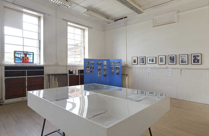 Photographs are displayed on the walls of a white room. In the centre there is a white table topped with glass.