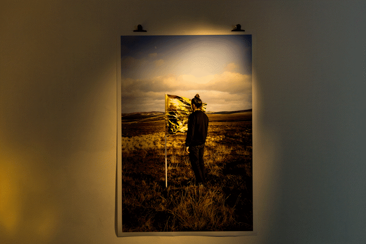 A print hangs on a white wall held by bullclips. It is in a dark room with golden light. The image shows a figure in black clothes, with brunette hair and topknot, standing with their face close to the fabric of a flag. The flag is made from a gold emergency blanket. The flag and figure are in an open field with hills in the far distance and blue sky.