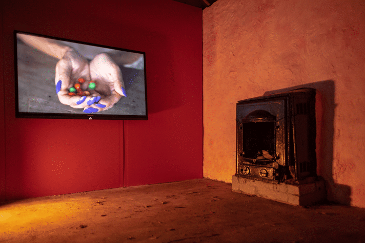 A room with orange walls and a soft light, with a dark fireplace on the right wall. There is a screen that depicts some hands put together as a bowl with purple painted nails holding colourful M&Ms.