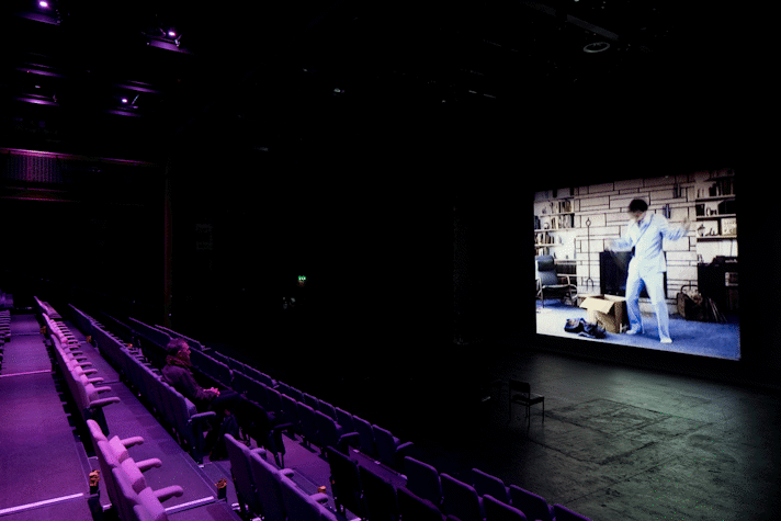 In a big dark room, there is a large projector screen on the right side and after a gap there are two chairs placed, facing the centre of the screen. Behind the chairs is seating for the audience, there are rows of permanent seating, of which there is a man sitting in the centre seat of the third row. The screen displays a man dressed in a suit making grand gestures, there is a box and a bag on the ground next to him. On the wall behind him there is a fireplace and bookshelves on either side.