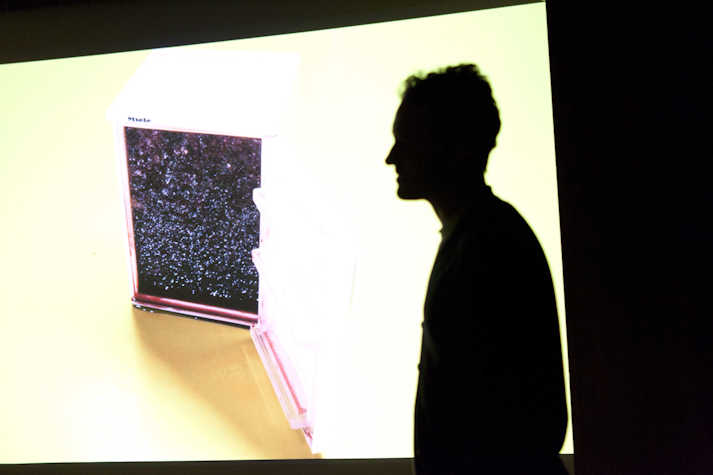 silhouette of a person in front of a large projection. the projection appears to show a small Miele fridge which is open revealing the night sky.