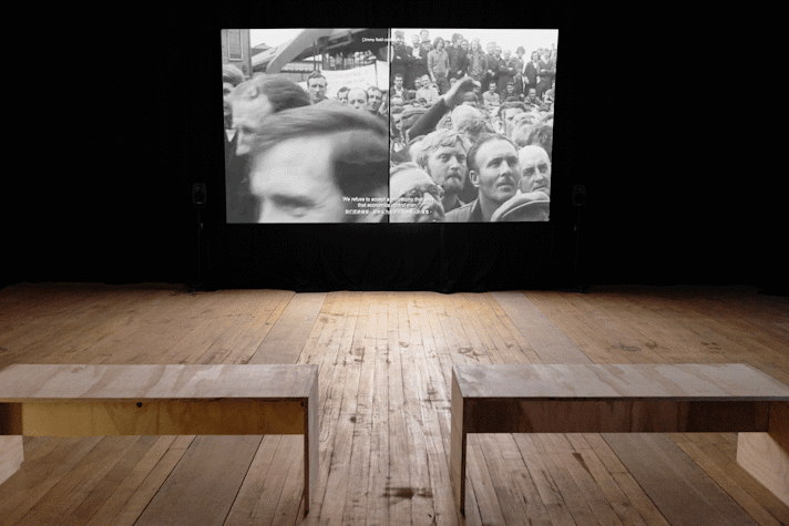 Film is projected onto a screen in a dark room. There are wooden benches facing the screen.