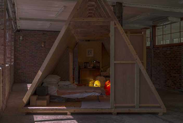 A triangular wooden structure in a open room with brick walls. Inside, there is light, and a bed can be seen.
