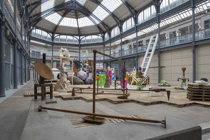In a large hall with a glass ceiling, there are multiple sculptures placed all around the room. Many are made of metal, stone and plastic.