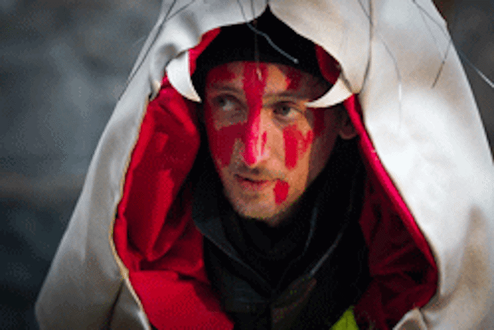 A photograph of a figure in a circus costume with a large white hood over their head and red paint on their face