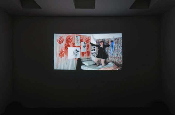 projection of film work onto a wall. it shows a woman standing on a bed in a bedroom. the bedroom walls are decorated with a large unstretched canvas of roses, with portrait drawings and paintings stuck on top.