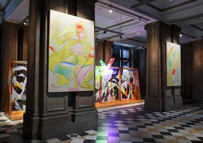 An abstract neon light showing two legs kicking the air in pointed heels, hangs at the top of a large wooden structure with glass panels and covered in fast gestural line paintings of figures and faces. The structure sits behind two stone columns on each of which a large scale canvas hangs, depicting fast gestural paintings of nude figures crouched and seated in pointed heels