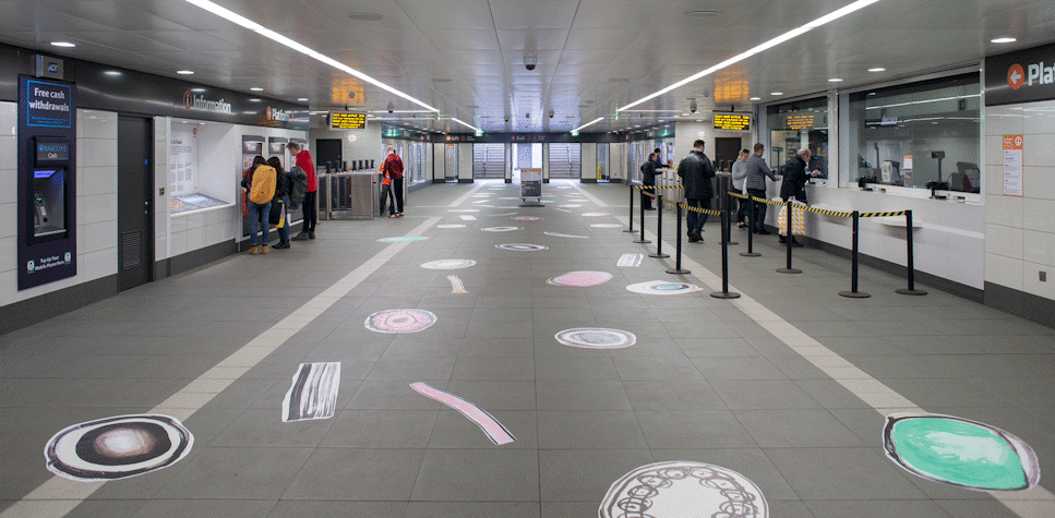 The subway entrance of St Enoch has circular drawn figures in the floor, in black, white, blue and pink colours. People are standing near the entrance and buying tickets.