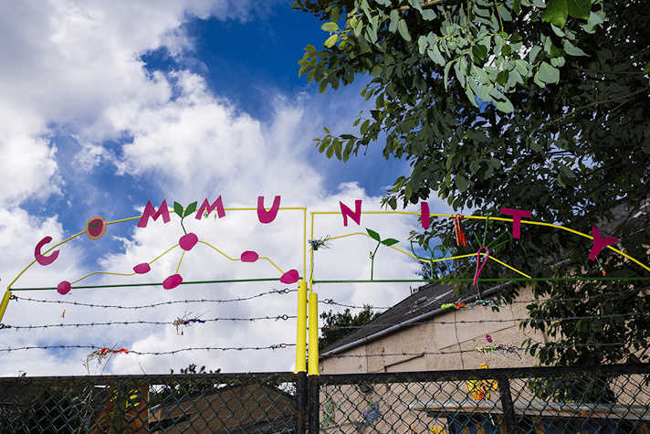 Above a gate the word 'Community' is spelled out in letters. Behind is a tree and clouds in the sky.