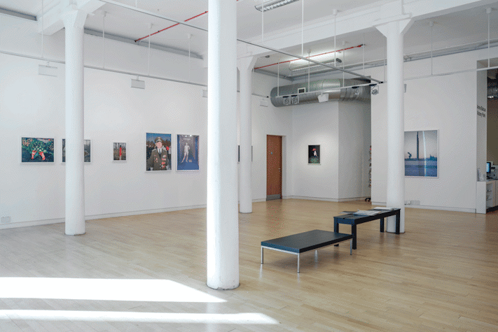 Photographs are centrally placed in a line along the walls of a large room. There are pillars standing across the room, with two low tables placed between two of the pillars.