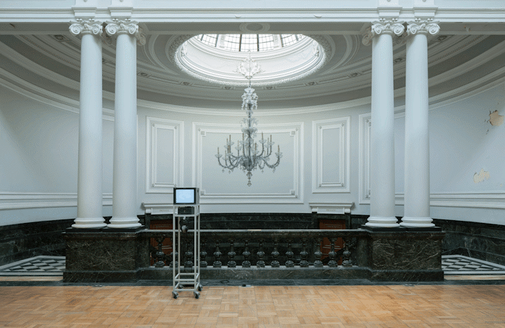 There is a room, with paint chipping off on the right side, a chandelier hanging from circular glass dome on the ceiling. There are two staircases going down on both sides, and nests to them on a marble pedestal are two poles on each side. The floor ahead of the poles is wooden and has a small stand with wheels, carrying small television monitor, with an image of the mathematical greater than and lesser than sign.