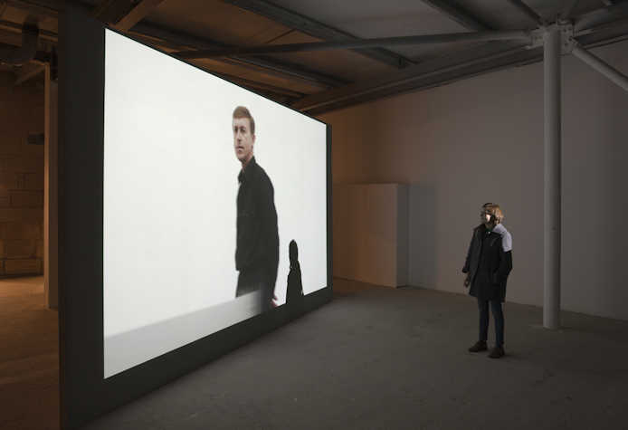 a person is stood in front of a large projection screen. on the screen there is a person looking off into the distance