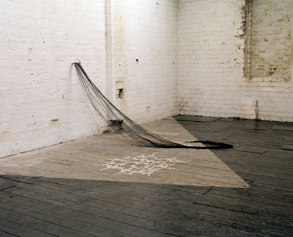 exhibition space with painted white brick walls. from the low on the wall hangs an installation of hair like thread that cascades onto the floor in a braid. next to it on the floor are small white beads arranged into a pattern.