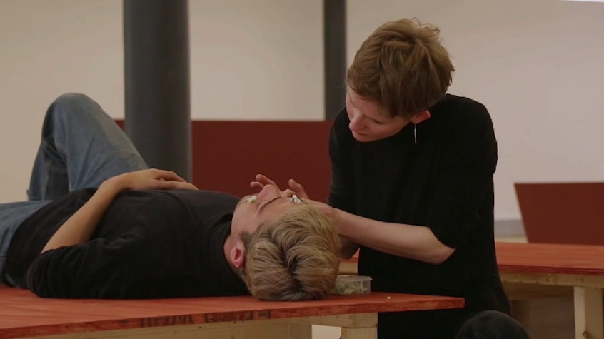 One blonde white person is lying down in a red table whilst the other person is putting flowers in their face. They are both wearing black sweaters.