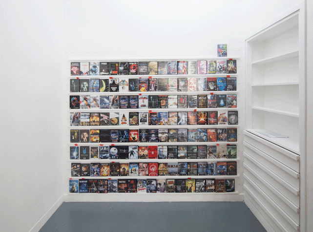 The wall is covered with rows of science fiction film DVD covers, Avatar and Total Recall among them. next to the wall is a perpendicular wall of shelves and chest of drawers in a minimalistic aesthetic corresponding to that of the wall.