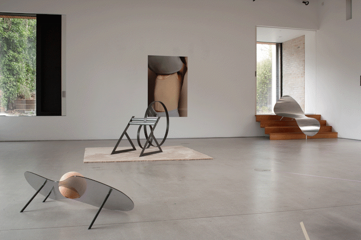 In a white studio, there are three different small sculptures made of metallic, plastic and tin materials. There is a photograph of someone taking off their sweater in the wall and two windows through which light comes into the room.