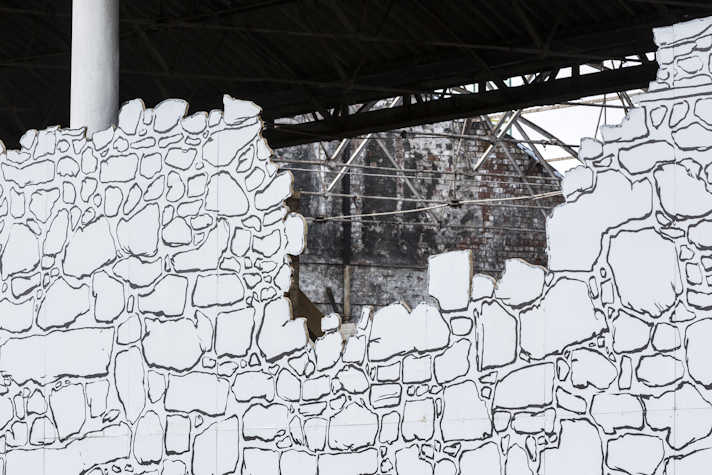 You can see an old building behind the black and white mural that resembles a stone wall. The mural is torn like a stone wall would be and allows the viewer to see the structure of the old building.
