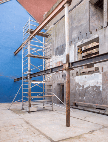 A patio surrounded by cement buildings. A blue wall in the background. Some tall metal scaffolding and a metal beam horizontally going through it.