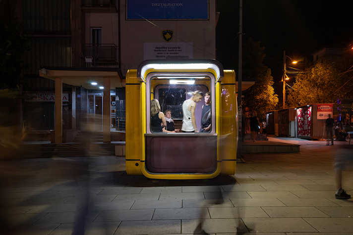Blurred images of people walk past a glowing yellow pod that sits on a street, there are people inside.