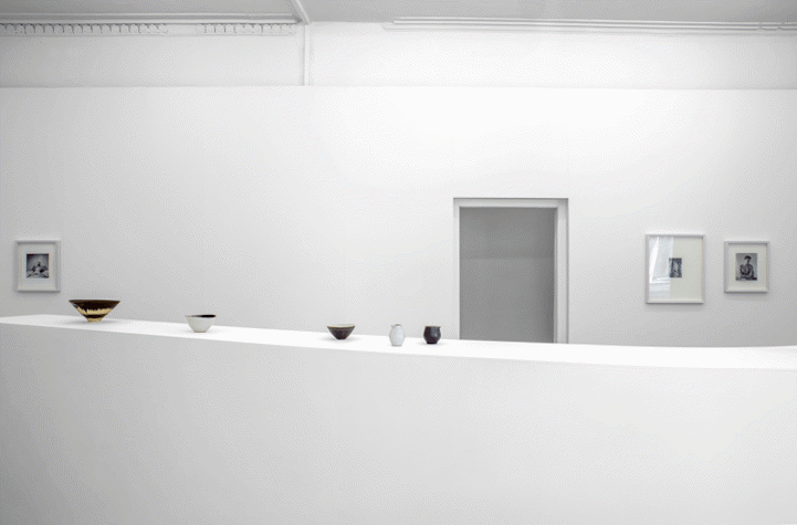 In a blank white room, there is a thin, long white table, on which there are multiple pottery pieces placed. They range in sized and colours. Behind the table, there is a door, with photographs hung on either side of it.