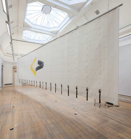In a long hall, with three glass domes on the ceiling, there is a long piece of fabric hung through the centre of the room. The fabric presents at the lesser than and greater than signs, facing each other and in different colours. The fabric has a rectangular grid like texture or print running throughput the fabric, with belts hanging off the bottom of each column.