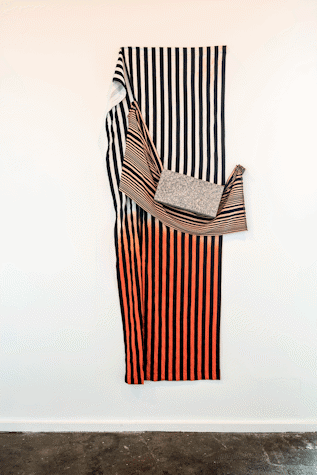 There is a cloth of black and white to red gradient lines that go vertically. In front there is a white and black horizontally stripped cloth holding a rectangular grey rock as part of Art installation in Tramway as part of Glasgow International 2018.
