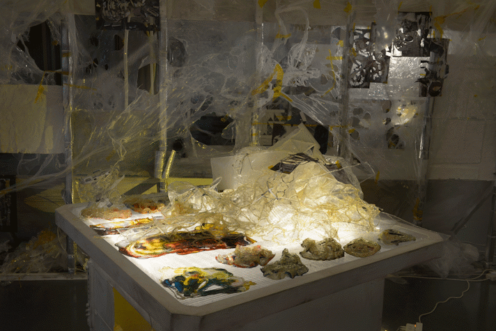 A table with light coming from within and different materials of different textures, colours and viscosity in web-like structures as part of an art installation in the Gallery of Modern Art, Glasgow International 2018.