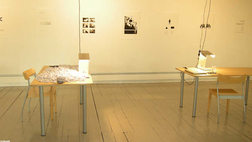 two tables with two chairs in a gallery space with images on the walls. both tables are covered in scrap paper.