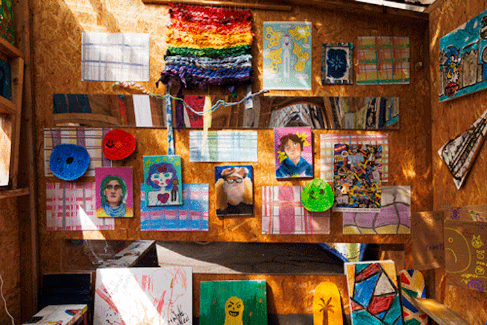 A collage of paintings and artwork are displayed on a wooden wall, with scattered sunlight coming through.