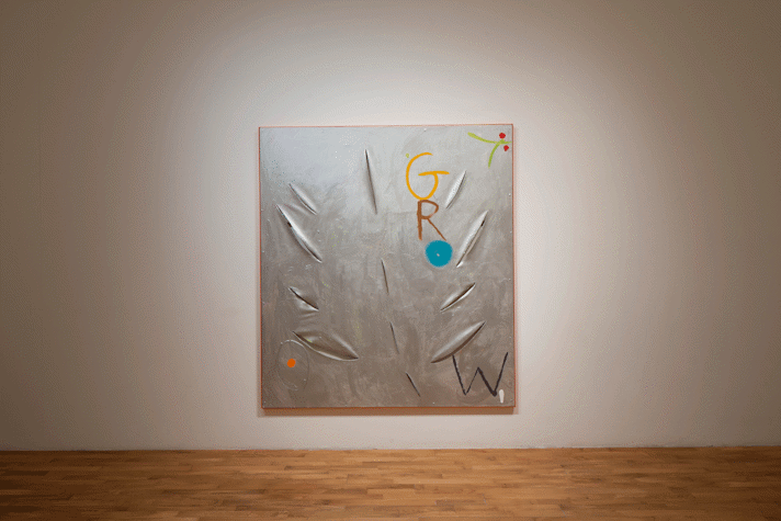 On a blank wall, hangs a large rectangular painting, with a light shining on it. The floor beneath it is wooden. The painting is metallic and has slashes in the shape of a leaf and the word "Grow" painted on it. On the right top corner is a peeking branch with a couple berries painted on it.