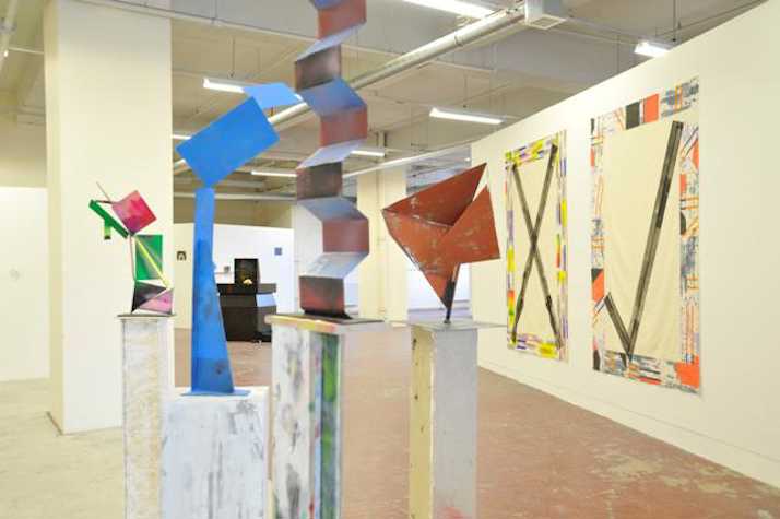 colourful geometric sculptures atop plinths. background shows two canvases, one with a cross and one with a tick, bordered by coloured patterns