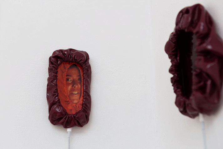 a painted red face in a red hoodie with the drawstring pulled tight looks out from an iphone mounted on the wall and on charge. the phone also has a drawstring hood around it.