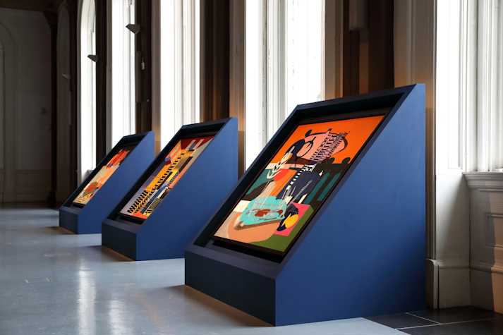 The image shows an exhibition of Nirbhai (Nep) Singh Sidhu's work in the Gallery of Modern art. The image shows three blue boxes holding colourful paintings.