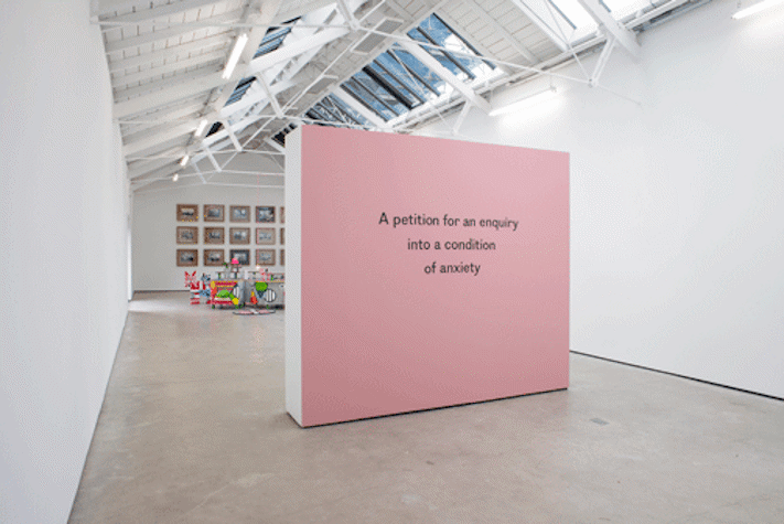 A white-walled gallery with a pink partition wall in the foreground. Multi-coloured sculptural works can be seen in the back ground, and a grid of photographs on the wall behind