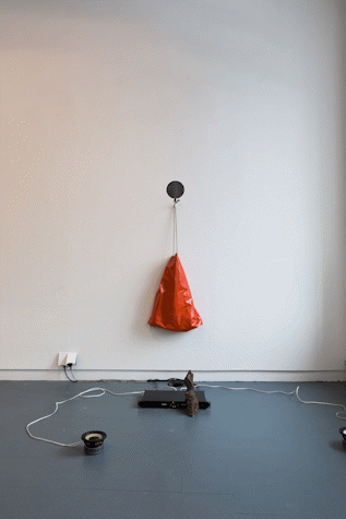 abstract sculptural assemblage on gallery wall and floor. Objects include an orange plastic drawstring bag, speaker, media player and block of wood