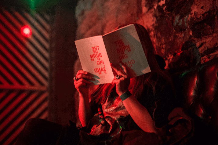 A person is sitting in the corner of a room which has dim red lighting. They are reading a book. The title of the book is "The Burning Sand".