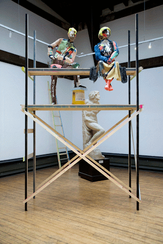 two colourfully painted and dressed humanoid sculptures sat atop scaffolding with a marble statue behind.