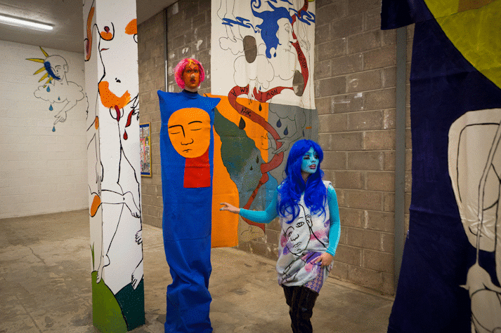 Playful and surreal line drawings of figures with blocks of colour cover a wall, column and fabric hanging in an industrial space. There are two performers, one appears to be extra tall and is wearing a tabard-like column of blue fabric with a disappointed looking cut out of an orange face, the performer's face is painted orange and they wear a red wig. The other performer has a turquoise painted face and electric blue wig, the line drawings of the wall cover their clothing.