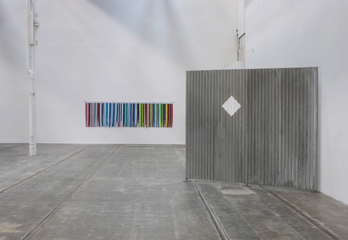In a big white room with grey floors a metal grey panel is to the right, with a diamond shape cut out of it. In the further wall, colourful strings are hanged (red, white, blue, green, yellow and black).