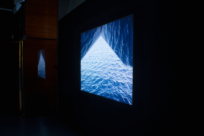 In a dark corner of a room, there is a projector screen placed on the wall with its image reflected on a glass door on its left side. The screen shows the same image, of ocean waves, with the image layered on top of itself, placed on the top left and right side of the backdrop of the same ocean.