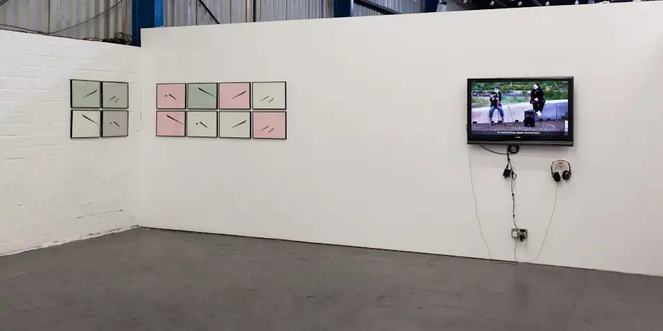 twelve black-framed A3 size paper works, on sage, grey, off-white and pink paper are hung in a grid of 4 and 8 on two adjacent walls. A small flat-screen TV plays a video with two sets of headphones hanging from it.