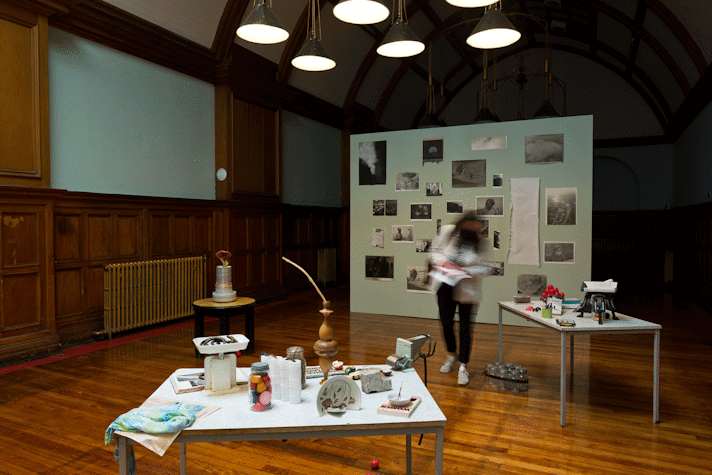 Lots of small objects sit on two tables, black and white images are displayed on a wall in the background