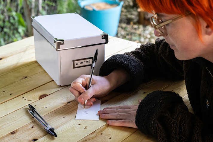 A person writes down something on a piece of paper leaning on a wooden table, next to a white box.