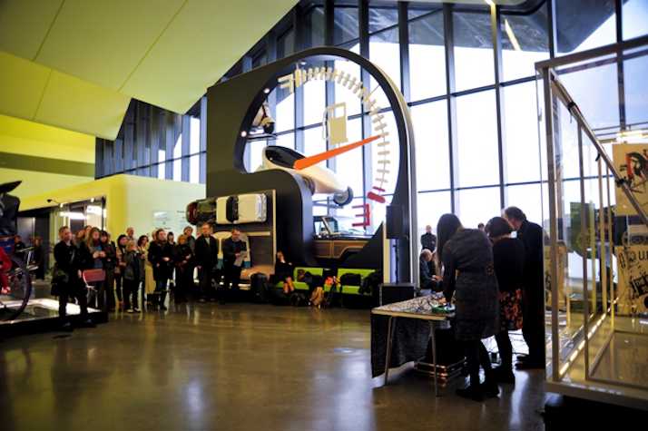 group of people convened in Glasgow's Riverside museum. A very large fuel gauge and model cars stand behind the crowd.