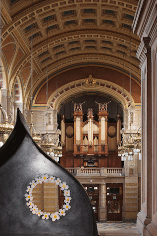 Black conical metal structure with a see-trough hole in it surrounded with a white flowers. The Kelvingrove Museum organ can be seen behind it.